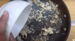 Pouring water into a sizzling pan of sautéed chopped onions on a wooden surface.